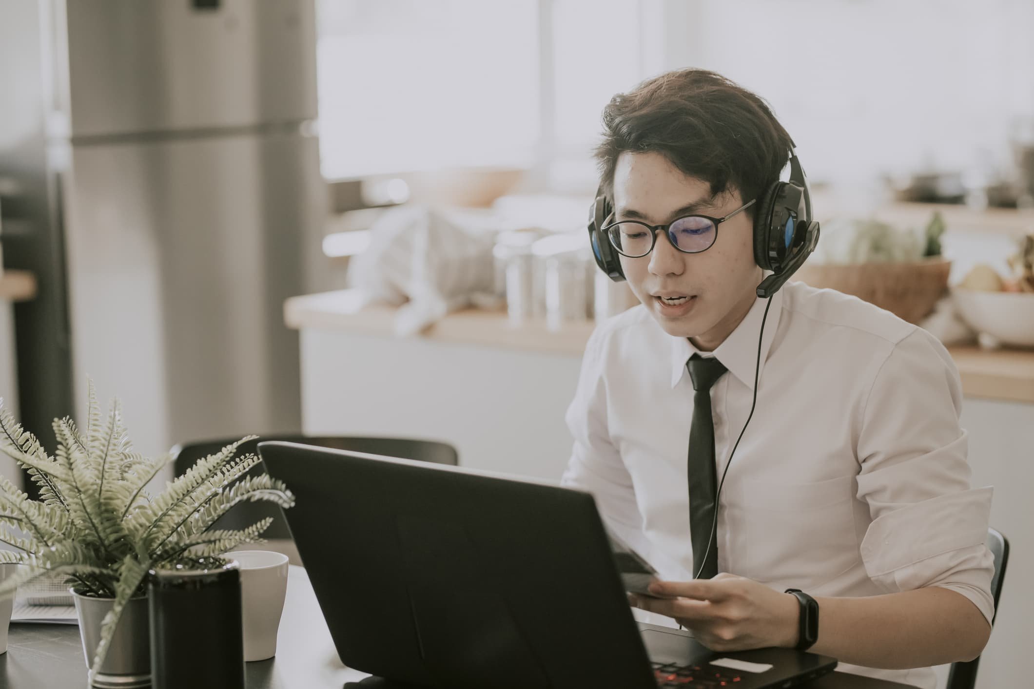 An asian chinese young adult man with tie, long sleeve shirt and short pant sitting using laptop in his dining room for video conference with his business partner and colleague virtual business meeting