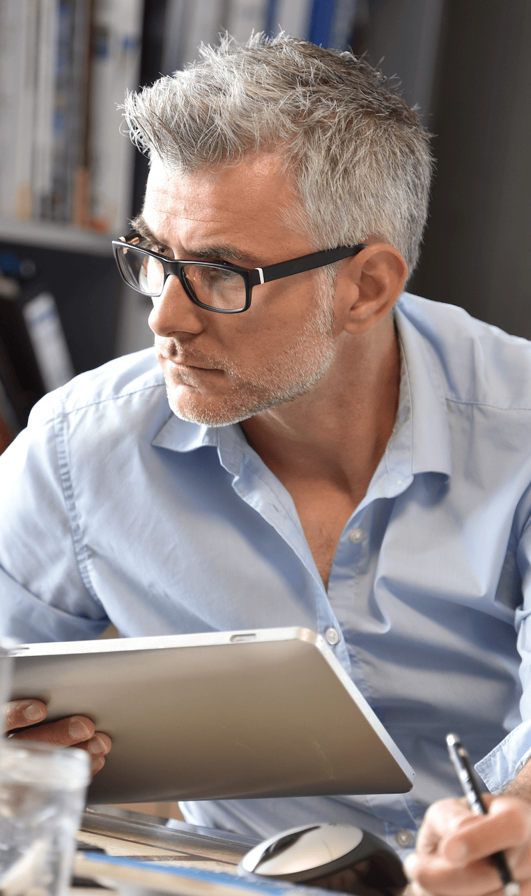 business-owner-mod-enterprise A man with gray hair and glasses is attentively looking to the side while holding a tablet, showcasing MyOutDesks enterprise solutions. Dressed in a light blue shirt, he sits at a desk, pen in hand, surrounded by various office items.