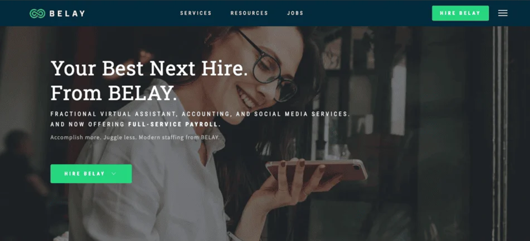 belay-ss1 A woman with glasses smiles while looking at her smartphone in a cafe. Text overlay highlights BELAY’s accounting and social media services, positioning them among the best virtual assistant companies, with a green Hire BELAY button.