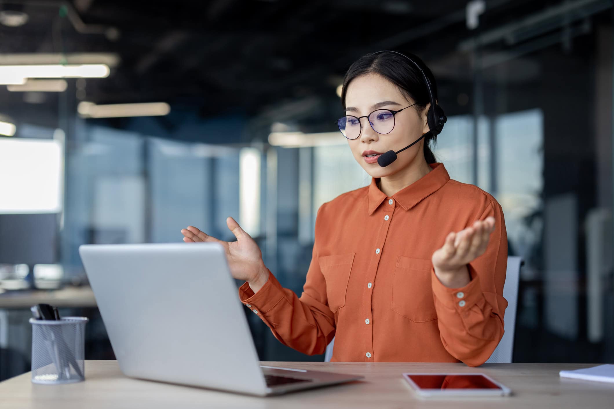 Serious asian woman with headset talking remotely using laptop for video call, help desk worker explaining to customers, business woman inside office at work, helpline worker Friendly businesswoman wearing a headset, smiling while assisting customers via video call on her laptop, working as a help desk professional in an office setting.