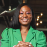 A person with shoulder-length hair smiles, wearing a bright green blazer. They have pink eyeshadow and light nail polish, seated with hands folded. The background features blurred lights, capturing the essence of professional headshots perfect for V-Taybrons portfolio.