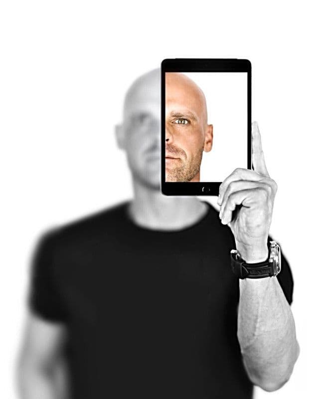 tony-giordano A person holds a tablet in front of their face, displaying half a face perfectly aligned with the other half visible, crafting an illusion. Against a white background and clad in black, they represent the seamless integration of real estate virtual assistant services into modern technology.