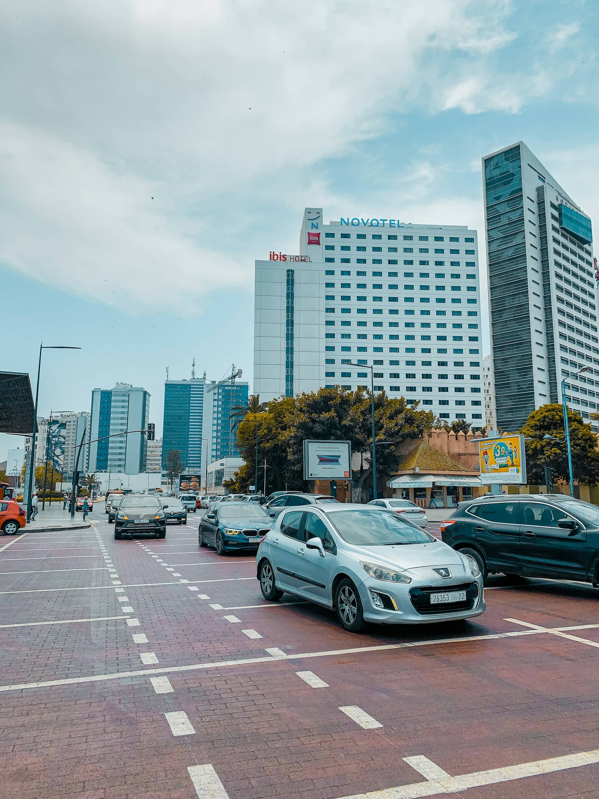 Cars wait at a traffic light on a city street lined with tall buildings, including the Ibis and Novotel hotels. The sky is partly cloudy, and some trees and billboards are visible along the road.