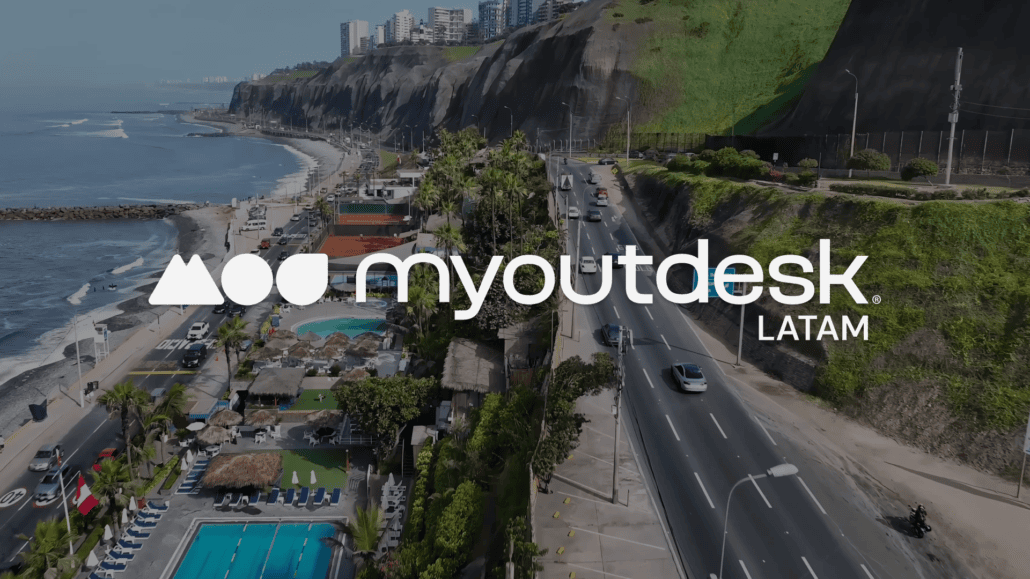 Aerial view of a coastal road and beach with pools and palm trees, city buildings on cliffs above, green hills, and the myoutdesk LATAM logo—highlighting access to top Spanish speaking talent.