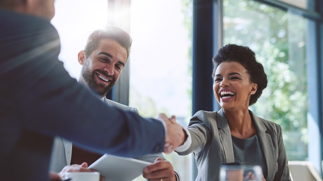 Two professionals, a man and a woman, smile and shake hands across a table during an outsourcing meeting in a bright, modern office. Another person is partially visible on the left.