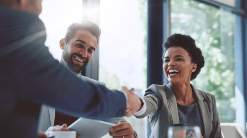 Two professionals, a man and a woman, smile and shake hands across a table during an outsourcing meeting in a bright, modern office. Another person is partially visible on the left.
