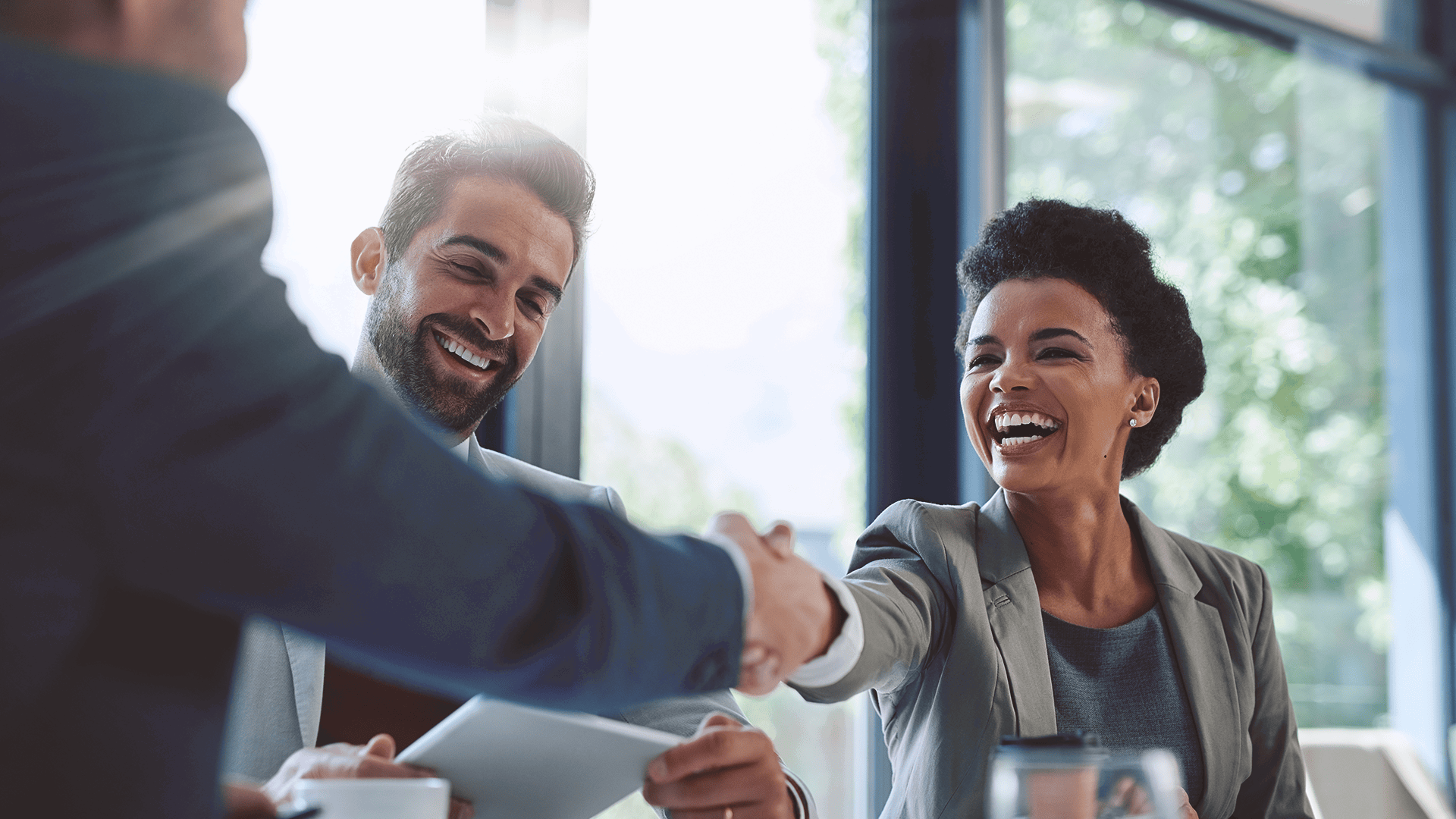 Two professionals, a man and a woman, smile and shake hands across a table during an outsourcing meeting in a bright, modern office. Another person is partially visible on the left.