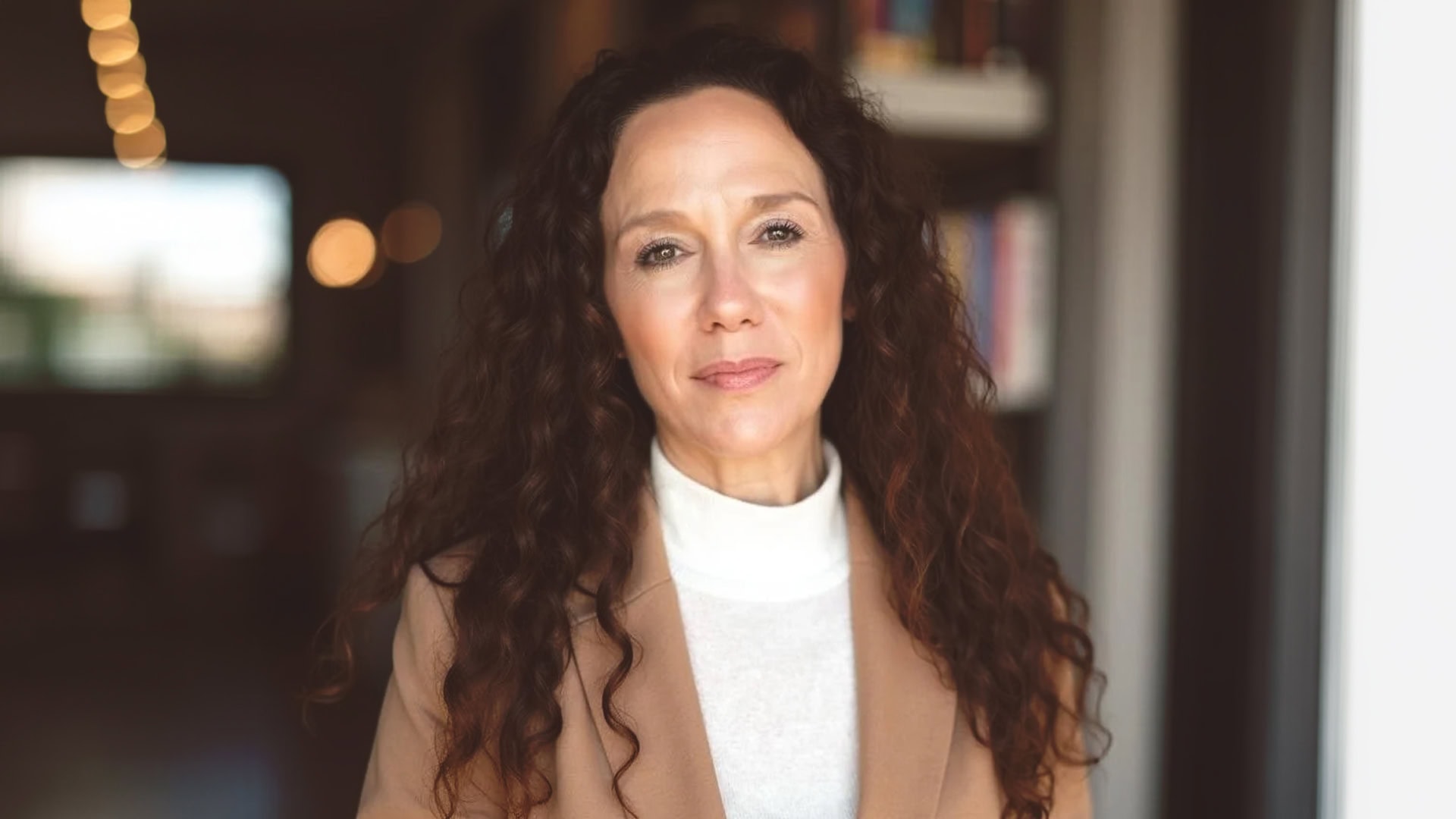 Erin Pedrami, a woman with long curly brown hair, wears a beige blazer over a white turtleneck as she stands indoors in soft lighting, shelves and blurred lights behind her. She has a neutral expression.