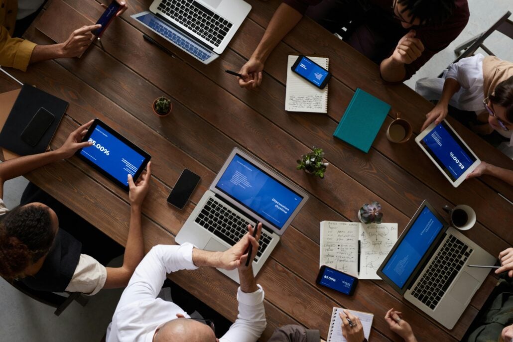 A group of people sit around a wooden table with laptops, tablets, notebooks, and phones, collaborating on a project. The devices display blue screens with charts and text as Spanish speaking talent contributes valuable insights.