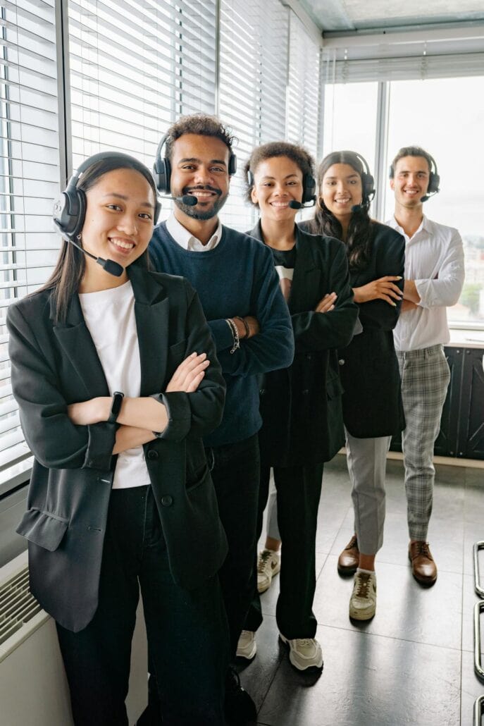 Five people stand in a bright office, four in business attire with headsets—reflecting an outsourcing team—smiling with arms crossed, while one in white casual clothes also smiles, large windows shining in the background.