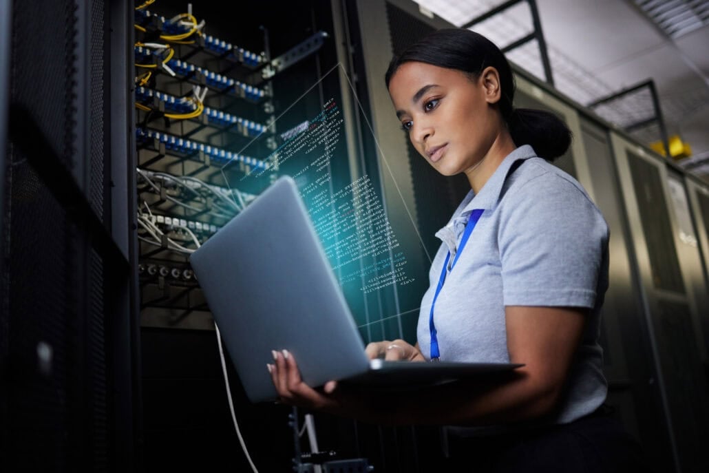 A woman stands in a server room, holding a laptop and working with code on the screen, showcasing tech talent from North Africa amid server racks and network cables.