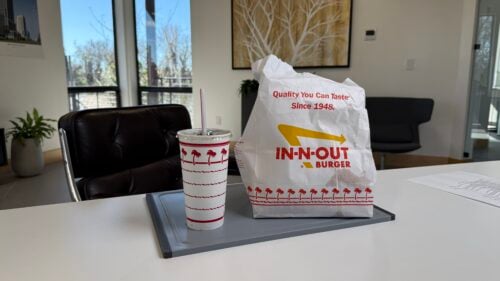 A drink cup and a paper bag with the IN-N-OUT Burger logo sit on a tray in a sunlit room, offering a perfect break for global distributed teams leadership brainstorming sessions among plants and modern decor.