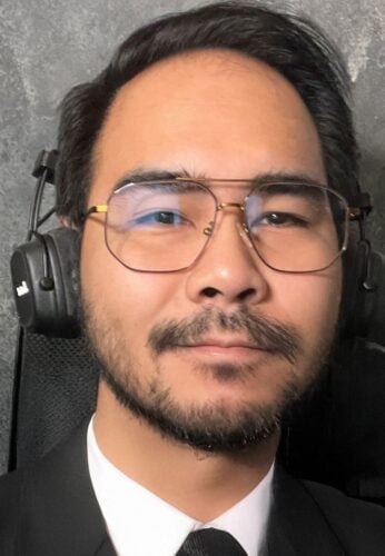 A man with glasses and a beard, wearing a suit and tie, sits in front of a gray textured background with black headphones over his ears, appearing to use the product as customers might in a professional setting.