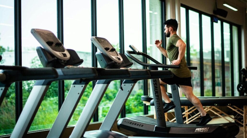 A man in athletic clothes runs on a treadmill inside a gym with large windows, letting in natural light and offering a view of greenery—a calm escape from scheduling chaos. Several treadmills are lined up in the room.